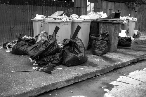 A row of five large wheelie bins positioned on a concrete pavement, with some overflowing with rubbish and additional black plastic garbage bags stacked in front. The bins are in varying shades of light grey and dark grey, with the lids mostly closed but some slightly ajar, revealing scattered paper and plastic waste. The surrounding environment includes a wooden fence in the background, indicating a residential or commercial area, and a section of the pavement shows signs of dirt and small debris. The overall scene suggests a rubbish collection or clearance effort, consistent with a waste removal service operating within the Stoke Newington area. The lighting appears natural and suggests daytime conditions, emphasizing the utilitarian aspect of rubbish disposal and clearance.
