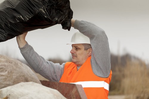 A waste management worker wearing a white safety helmet and an orange high-visibility vest is seen lifting a large black rubbish bag with both hands, preparing to load it onto a pile of similar refuse. The background features an outdoor setting with an overcast sky, sparse vegetation, and a distant view of blurred trees or shrubs. The worker's focused expression indicates careful handling of waste materials. In the foreground, there are rough-textured, light-colored bags or bundles, possibly filled with waste, placed on what appears to be a paved or gravel surface. The scene is part of a rubbish removal operation, likely conducted by Stoke Newington House Clearance, serving the local area around N16 postcode, and illustrates proper safety gear and loading procedures in a professional rubbish collection context.