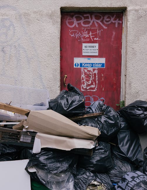 Piles of black rubbish bags, some partially torn or collapsed, are stacked in front of a red metal door set into a beige, textured exterior wall. The bags appear to contain waste and are arranged haph